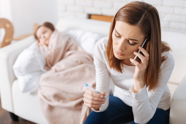 Cheerless troubled mother holding thermometer and calling a doctor while her ill daughter sleeping in the background