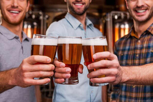 乾杯！金属製の容器の前に立っている間、ビールとメガネを伸ばして笑顔でカジュアルな服装で3人の陽気な若い男性のクローズアップ