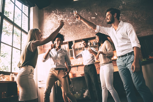 Cheers to friends! Low angle view of cheerful young people dancing and drinking while enjoying home party on the kitchen