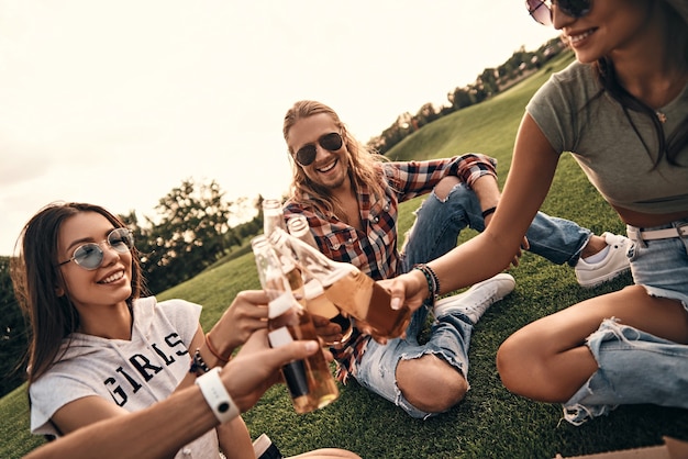 Cheers to friendship! Group of young smiling people in casual wear toasting with beer bottles while enjoying picnic outdoors