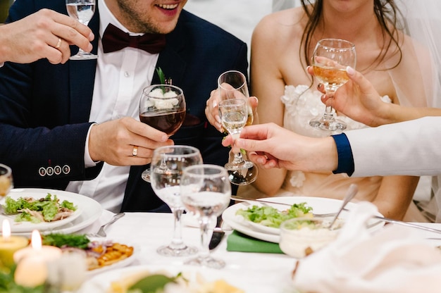 Cheers Newlyweds with friends drink champagne celebrate at the table People raise glasses of wine and vodka for toast Group of men and women celebrate wedding