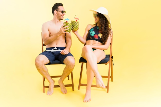 Cheers to our lovely honeymoon. Attractive couple of newlyweds in swimsuits sitting on beach chairs and making a toast with their pina colada cocktails