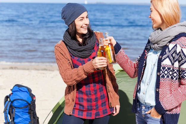 乾杯！ビーチでテントの近くに立っている間、ビールで応援し、笑顔で2人の美しい若い女性