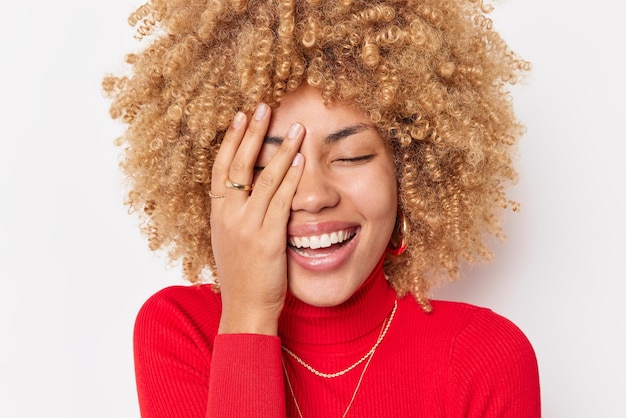 Cheerul European woman with curly blonde hair makes face palm gigles happily wears casual red turtleneck poses against white background. Laughter joy good joke and emotional reaction concept