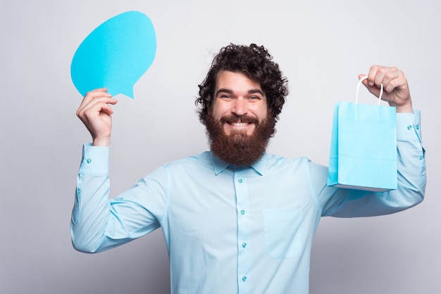 Cheerul young man in blue shirt holding speech bubble and shopping bag.