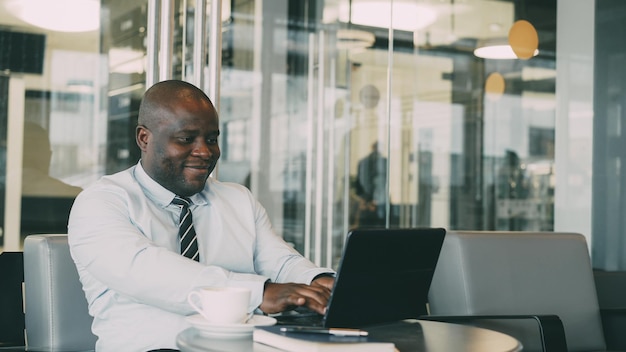 Cheery African American businessman smiling printing and working on his laptop in glassy cafe during lunch break