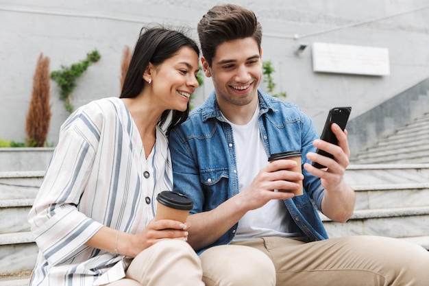 cheery couple man and woman in casual clothes drinking takeaway coffee and using smartphone on city stairs outdoors