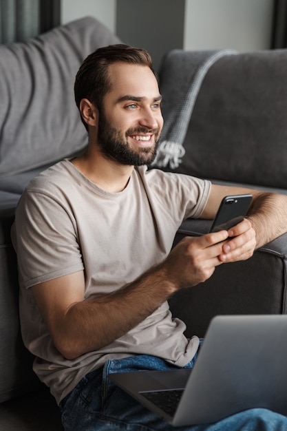 a cheery happy young man indoors at home on sofa using laptop computer chatting by mobile phone.