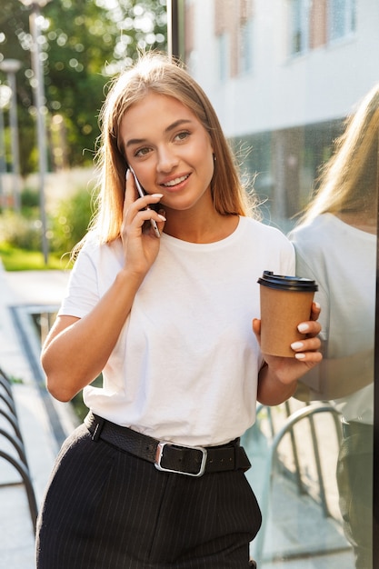 cheery pleased young business lady with cup of coffee posing outdoors near business center talking by mobile phone.