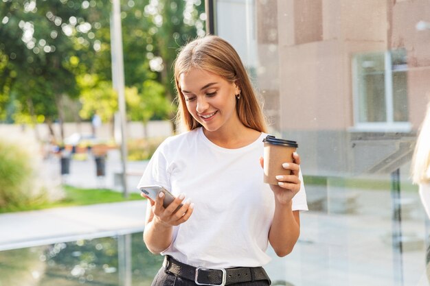 cheery positive young business lady with cup of coffee posing outdoors near business center using mobile phone.