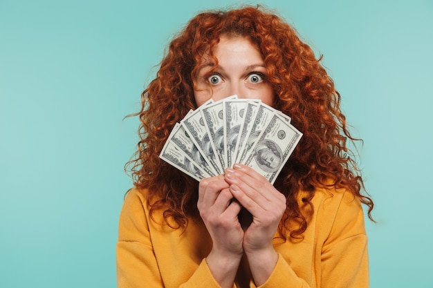 cheery redhead woman 20s smiling and holding bunch of dollars money isolated over blue wall