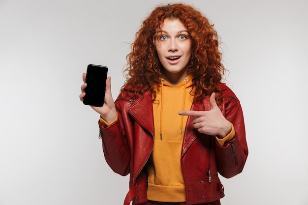 白い壁に隔離されたスマートフォンを笑顔で保持している革のジャケットを着ている陽気な赤毛の女性20代