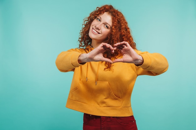 Photo cheery redhead woman 20s wearing leather jacket smiling and showing heart shape with fingers isolated over blue wall