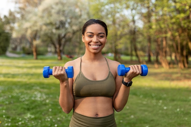 Cheery young black woman doing outdoor strength training\
exercising with dumbbells working out