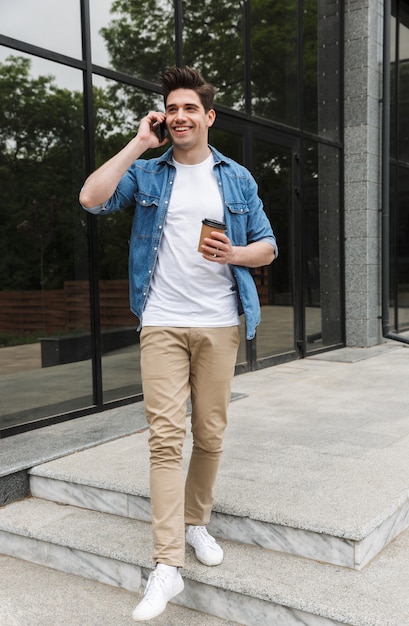 cheery young man in casual clothes drinking takeaway coffee and talking on cellphone while standing over building