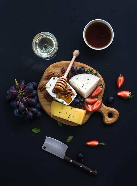 Cheese appetizer selection or whine snack set Variety of cheese grapes pecan nuts strawberry and honey on round wooden board over black backdrop