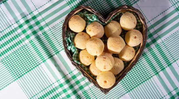 Cheese bread, heart-shaped basket lined with green and white fabric filled with cheese bread on a checkered tablecloth, top view.