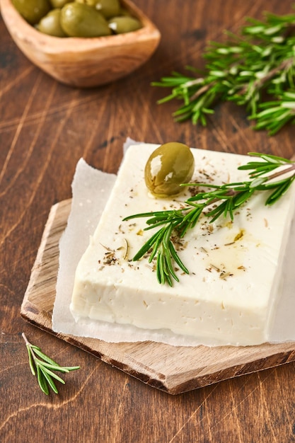 Feta di formaggio con rosmarino, erbe aromatiche, olive e olio d'oliva su tagliere di legno su fondo di legno vecchio. formaggio casalingo greco tradizionale. messa a fuoco selettiva.