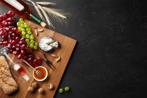 Cheese head, bunch of grapes, honey, nuts and wineglass on wooden board and black background. Top view with copy space.