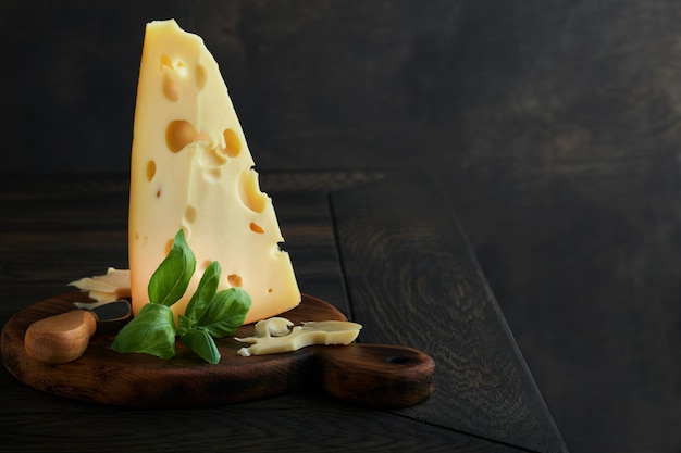 Cheese piece of cheese triangle swiss cheese on with basil and\
a knife on a serving board on dark wooden table background cheese\
background