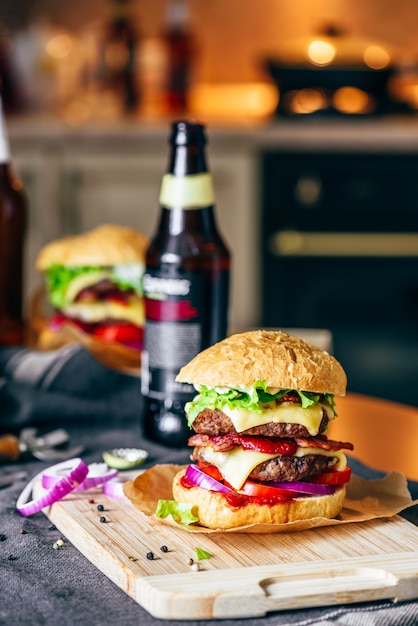 Cheeseburger con due polpette di manzo, formaggio cheddar, pancetta, lattuga iceberg, pomodori a fette e cipolla rossa. bottiglia di birra e alcuni ingredienti sul tavolo.