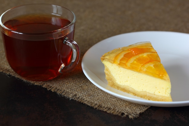 Cheesecake met sinaasappelen op een witte plaat, glazen mok met thee op jute tafelkleed.