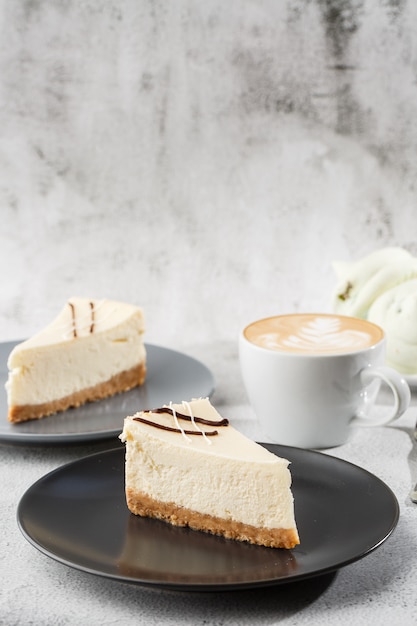 Cheesecake New York or classic cheesecake with cup of coffee on white table. Closeup view. Tasty breakfast. Piece of cake on black plate, white cup on white marble background. Vertical photo.