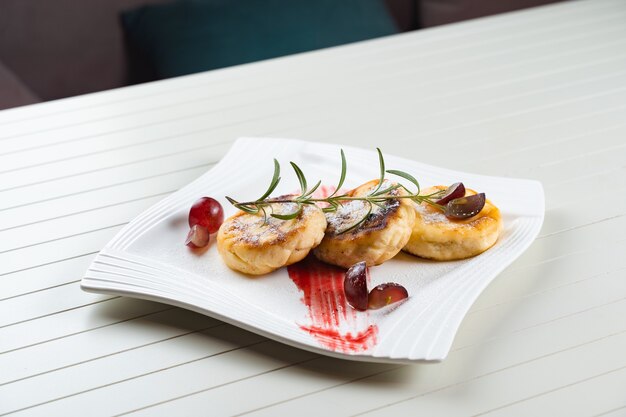 Cheesecakes decorated with powdered sugar, rosemary branch and grapes with berry jam on a white plate. Delicious cottage cheese breakfast.