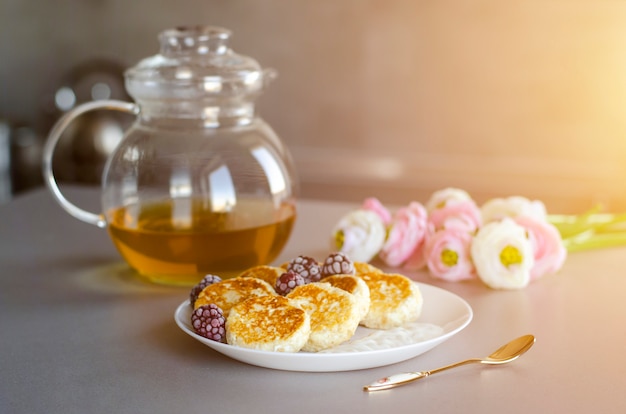 Cheesecakes from cottage cheese with blackberries on gray table with transparent teapot, tea spoon and flowers 