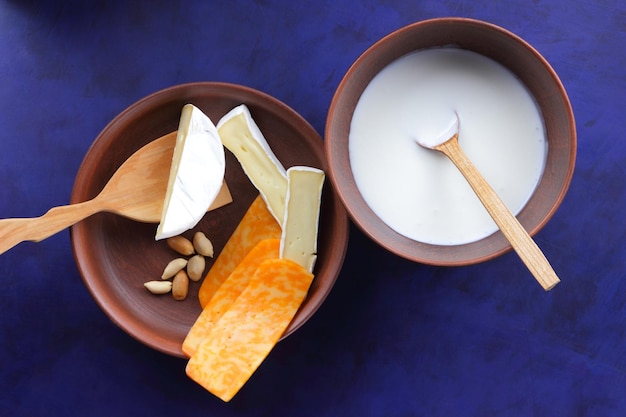 Cheeses with nuts and a wooden spatula on a clay plate Different types of cheese plate and a bowl of milk on a blue background closeup