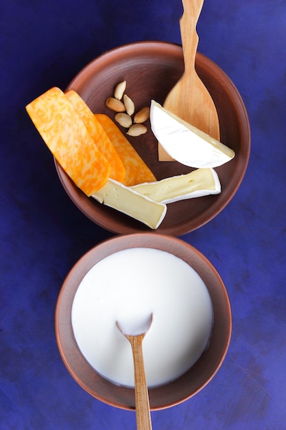 Cheeses with nuts and a wooden spatula on a clay plate Different types of cheese plate and a bowl of milk on a blue background closeup