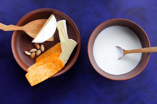 Cheeses with nuts and a wooden spatula on a clay plate Different types of cheese plate and a bowl of milk on a blue background closeup