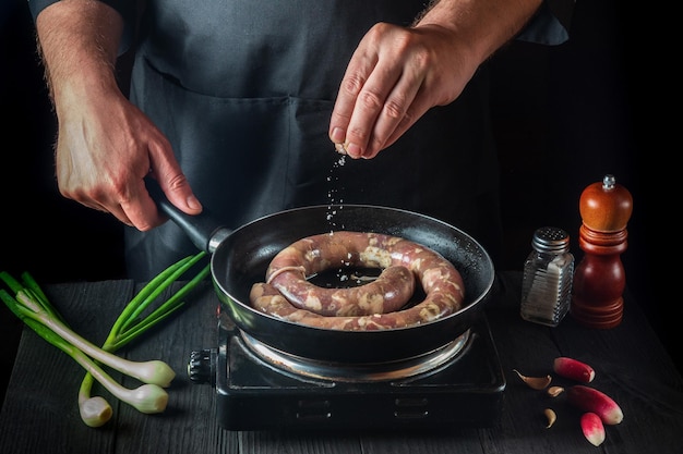 The chef adds salt to pan with raw meat sausage. Preparation for cooking sausages in the kitchen of a restaurant or cafe on table with vegetables and spices.