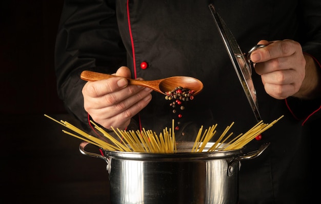 シェフは、スパゲッティの沸騰した鍋にスプーン一杯のコショウを加えます暗い背景にレシピまたはメニュー用の空きスペース