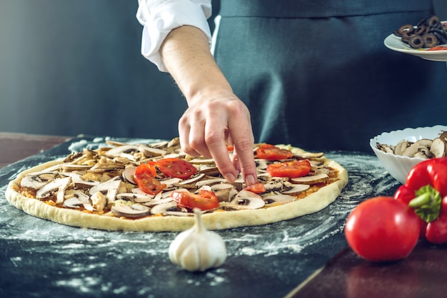 Chef in grembiule nero che mette i pomodori su una pizza