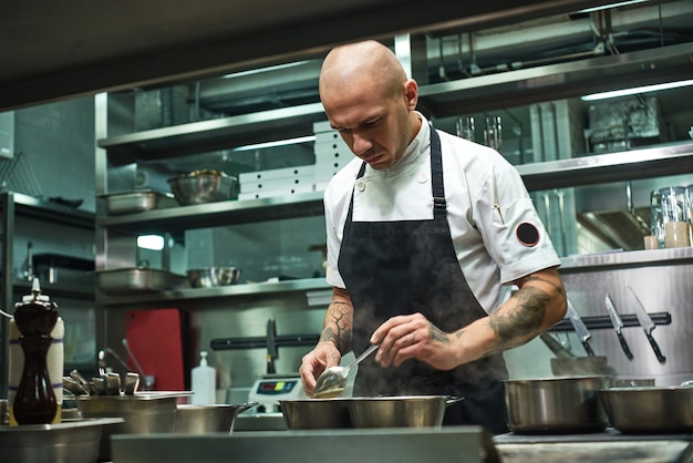 Chef in grembiule nero e con diversi tatuaggi sulle braccia che cucinano un piatto nella cucina del suo ristorante