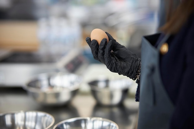 a chef in black gloves holds a chicken egg in his hand against the background of kitchen utensils