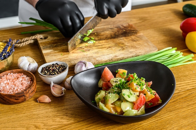 A chef in black gloves is slicing fresh green onions on a wooden chopping Board.