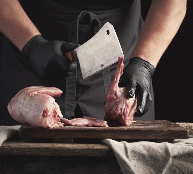 검은 라텍스 장갑을 입은 요리사가 큰 칼을 들고 생 토끼 고기 조각으로 잘라냅니다.