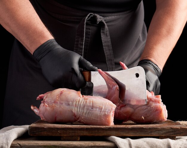 Chef in black latex gloves holds a big knife and cuts into pieces raw rabbit meat