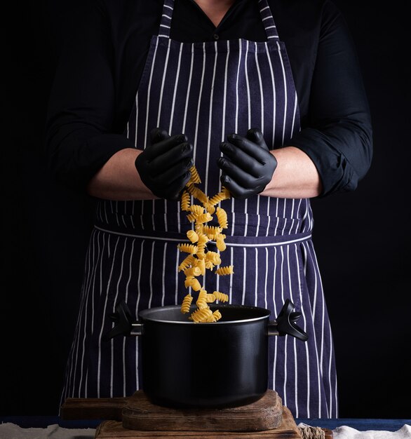 Chef in guanti di lattice nero, grembiule a strisce versa la pasta fusilli cruda in una padella di metallo