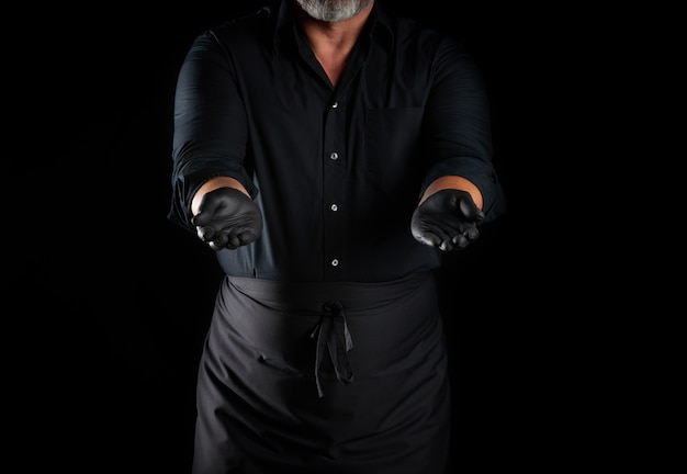 Chef in a black shirt, apron and latex black gloves stands on a black space, his arms are outstretched forward, holding an object