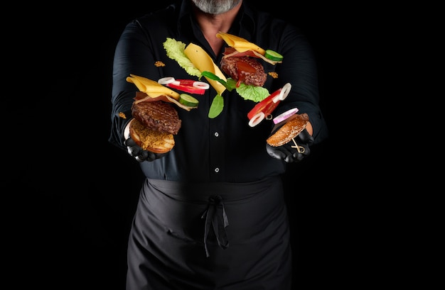 Шеф-повар в черной рубашке, фартуке и черных латексных перчатках стоит на черном пространстве, в его руках летают ингредиенты чизбургера: булочка с кунжутом, котлета, помидор, салатные и луковые кольца, сыр