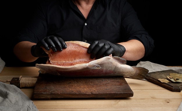 Chef in a black shirt and black latex gloves holds a raw carcass of headless salmon fish