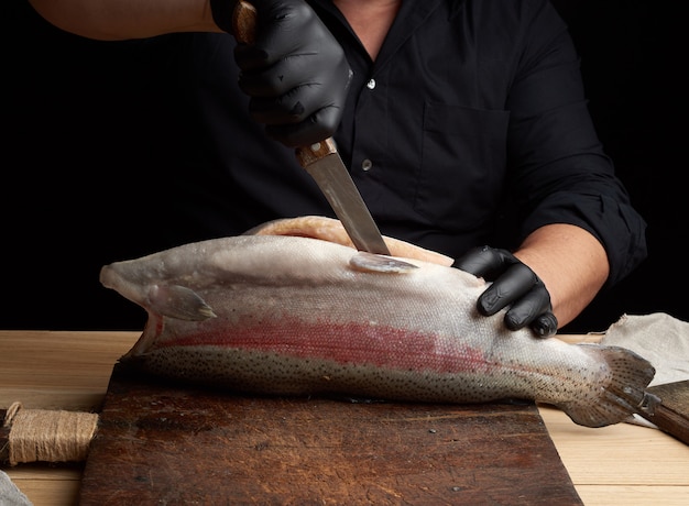 Chef in black shirt and black latex gloves slices a whole fresh fish salmon