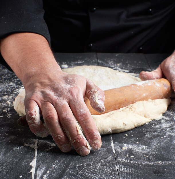 chef in a black tunic rolls a dough for a round pizza on a black table with a wooden rolling pin