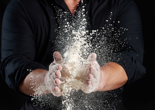 Foto lo chef in uniforme nera spruzza farina di grano bianco in direzioni diverse, il prodotto sparge polvere, sfondo nero, da vicino