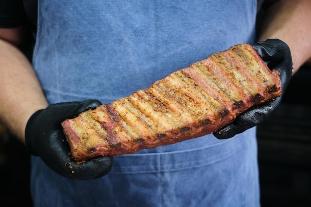 青いエプロンを着たシェフがグリルで焼いた肋骨のラックを握っている