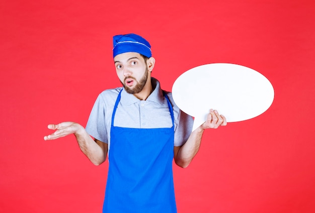 Chef in blue apron holding an ovale info board and looks confused. 