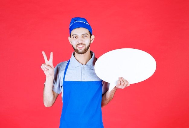 Chef in blue apron holding an ovale info board and showing enjoyment sign. 